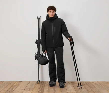 Un homme en tenue de ski noire tenant des bâtons, avec des skis et un casque à côté de lui.