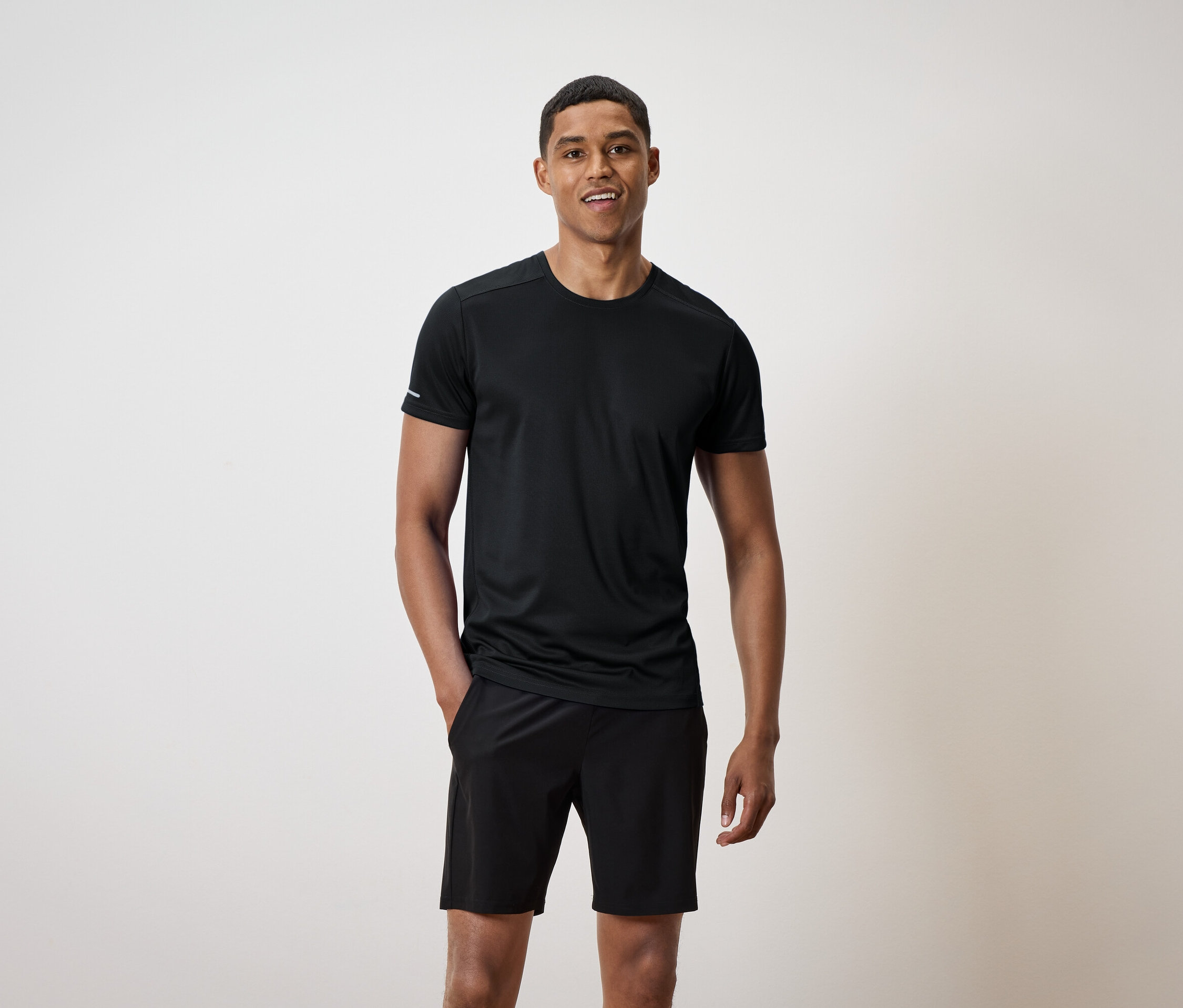 Un homme souriant se tient debout, les mains dans les poches de son short noir, et porte un t-shirt noir.