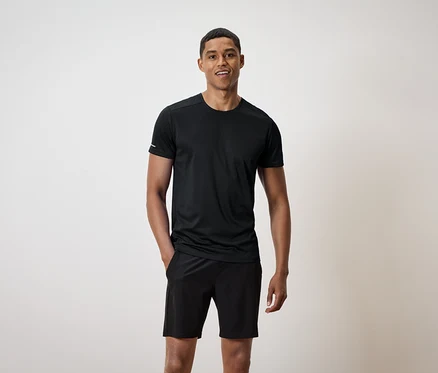 Un homme souriant se tient debout, les mains dans les poches de son short noir, et porte un t-shirt noir.