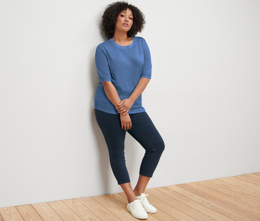 Une femme appuyée contre un mur portant un pull en maille fine bleu, un pantalon bleu marine et des baskets blanches.