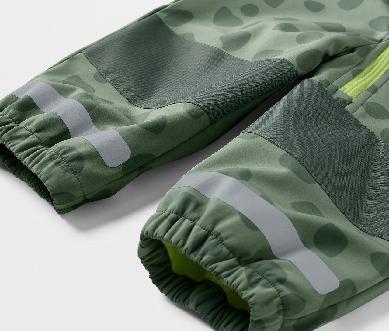 Gros plan sur une combinaison en softshell pour enfant avec un motif camouflage vert, des bandes réfléchissantes et des poignets élastiques.