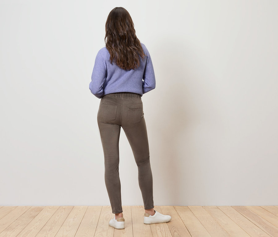 Une femme aux cheveux bruns ondulés se tient dos à nous, portant un sweat-shirt violet, un pantalon marron et des baskets blanches.