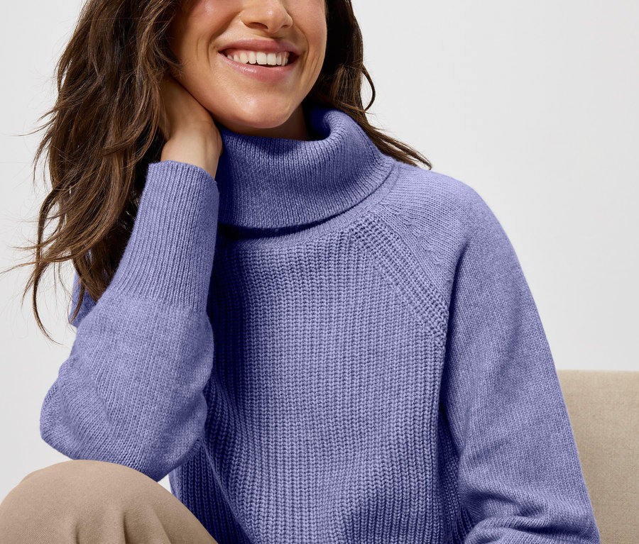 Une femme souriante est assise, la main sur la nuque. Elle porte un pull à col roulé violet et un pantalon beige.
