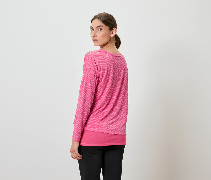 Une femme regarde par-dessus son épaule, portant un haut rose à manches longues et un pantalon noir.