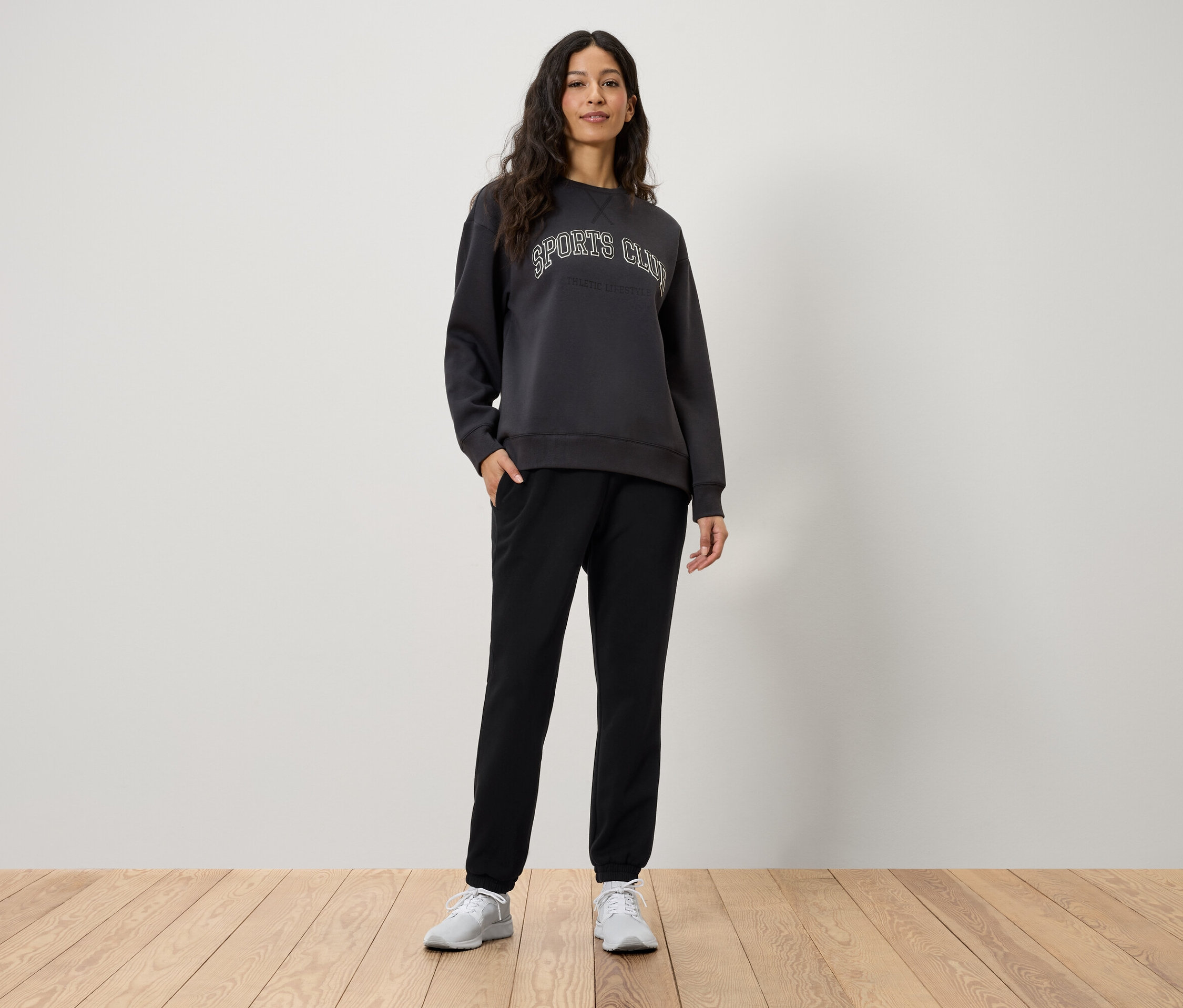 Une femme se tient debout, les mains dans les poches de son pantalon de jogging noir. Elle porte un sweat-shirt gris avec l'inscription Sports Club et des baskets blanches.