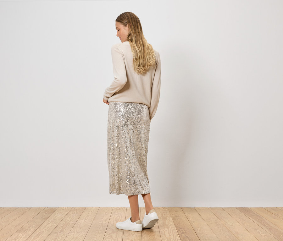 Femme posant en pull beige et jupe à paillettes avec des chaussures argentées.