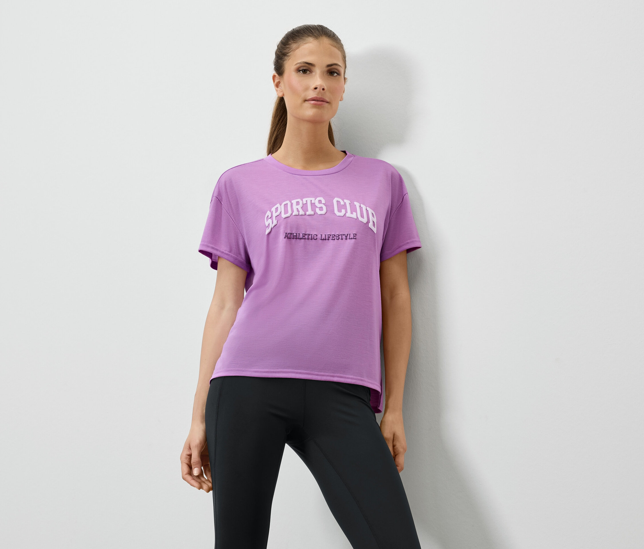 Une femme portant un t-shirt lilas avec un imprimé et des leggings noirs pose devant un mur blanc.
