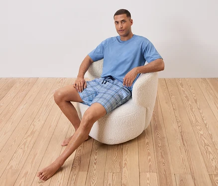 Un homme est assis pieds nus dans un fauteuil blanc. Il porte un t-shirt bleu clair et un short à carreaux bleus.