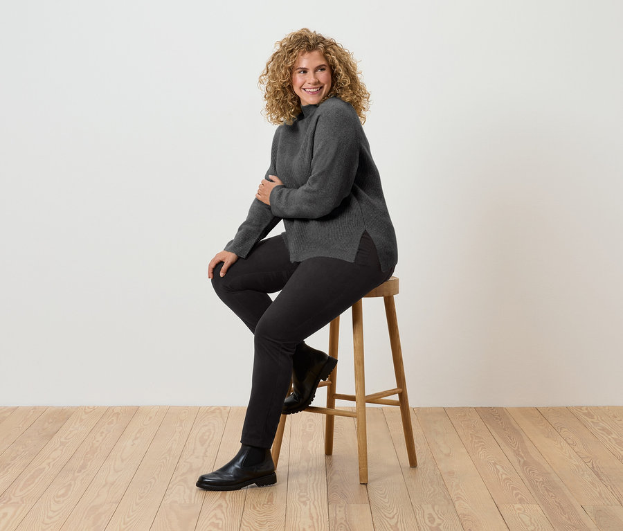 Une femme aux cheveux bouclés est assise sur un tabouret en bois. Elle porte un pull à col roulé gris, un pantalon noir et des bottes noires.