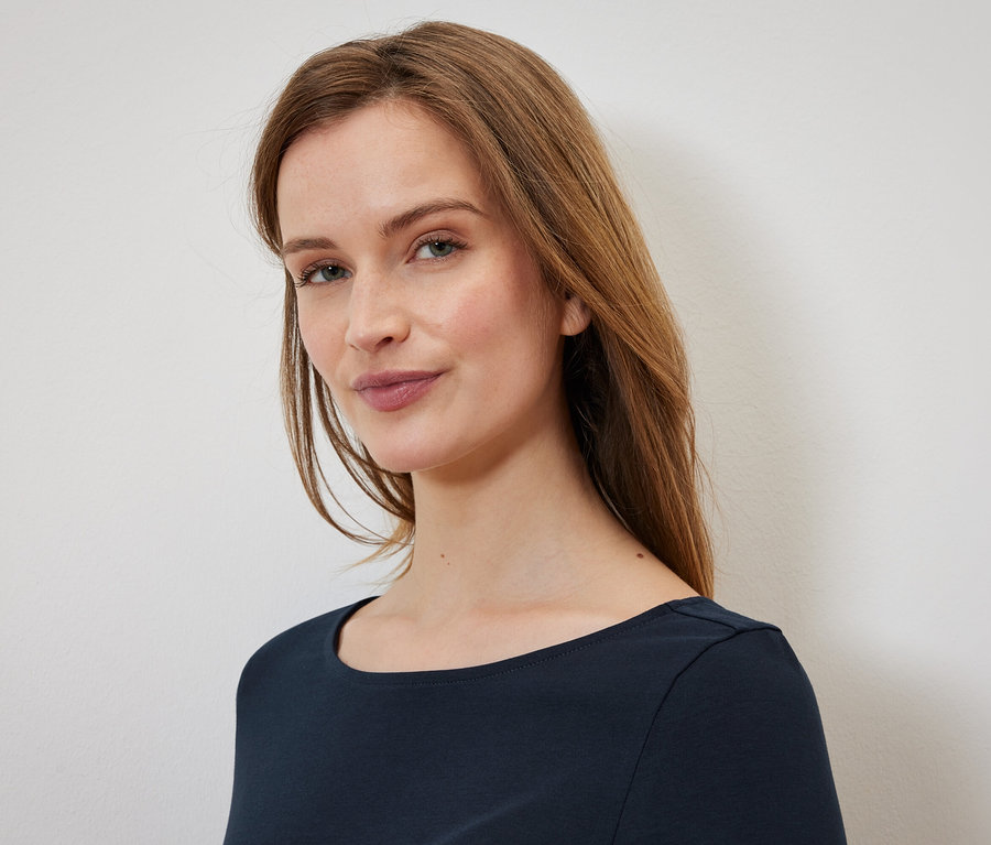 Portrait d'une femme aux longs cheveux bruns et aux yeux bleus, portant un t-shirt bleu foncé.