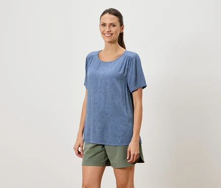 Une femme avec une queue de cheval porte un t-shirt bleu à manches courtes et un short vert sur un fond blanc.
