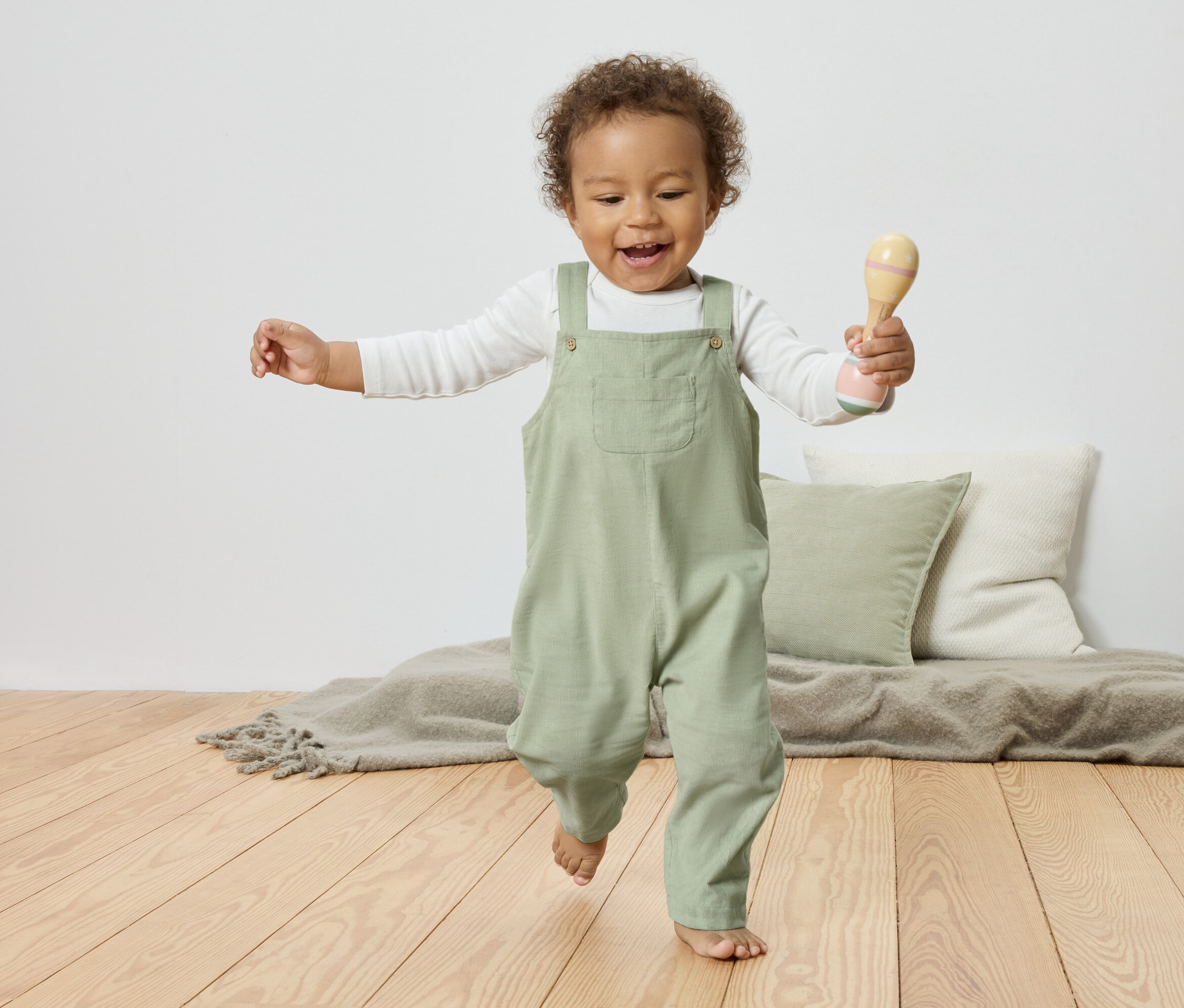 Un enfant souriant aux cheveux bouclés, vêtu d'une salopette vert clair, tient un hochet à la main et court sur un plancher en bois.