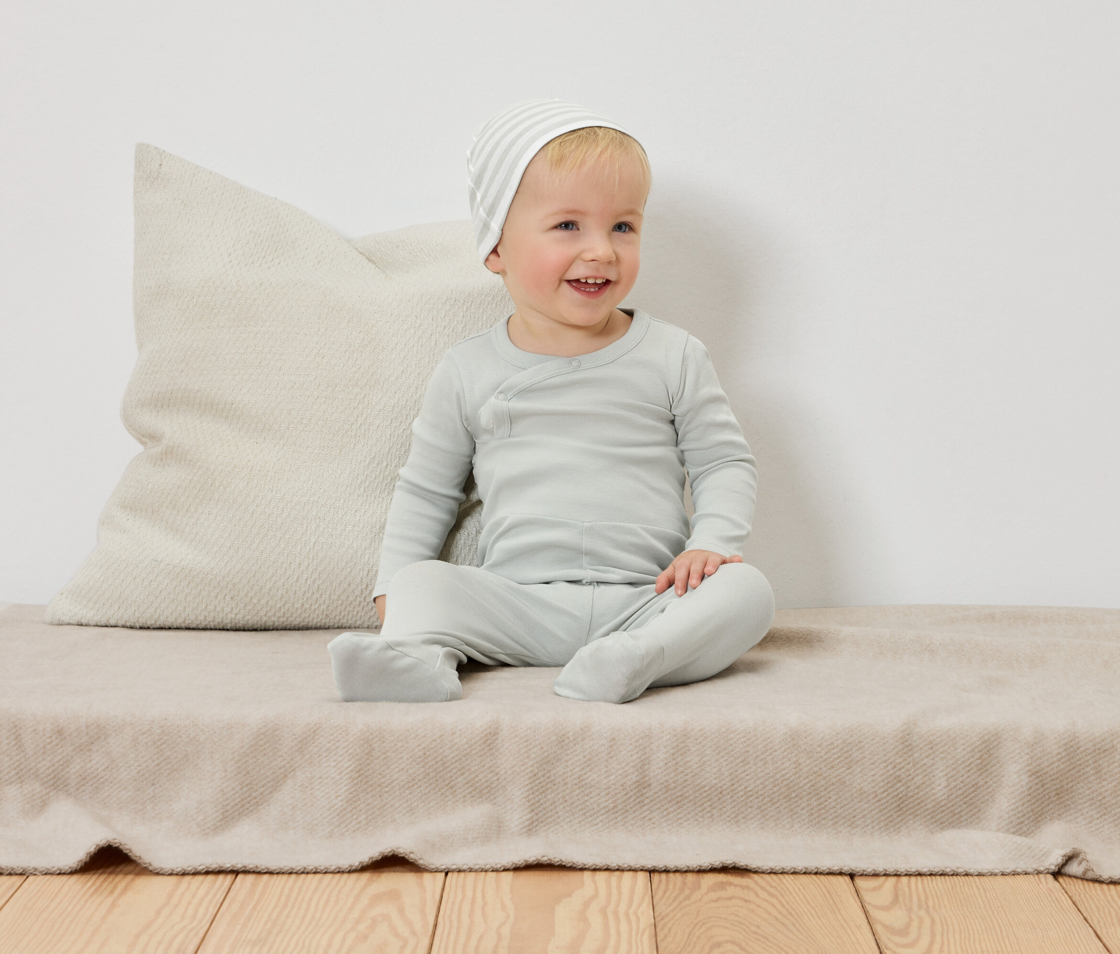 Un bébé souriant avec un bonnet rayé est assis sur une couverture beige devant un oreiller.