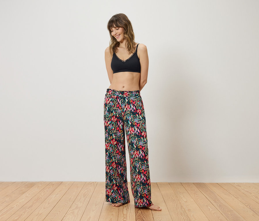 Femme en bralette noire et pantalon à fleurs debout devant un mur blanc.