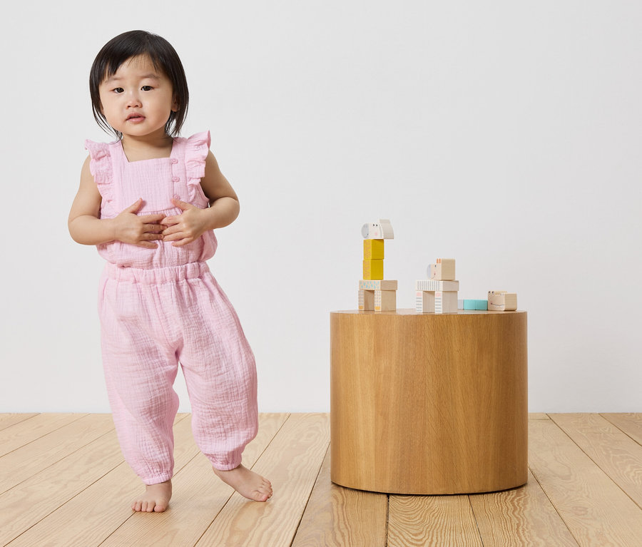 Petite fille en combinaison de mousseline rose debout à côté d'une table ronde en bois avec des blocs de construction.