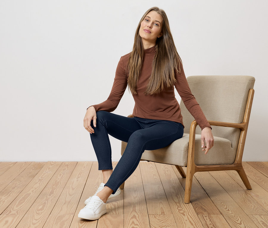 Une femme en haut marron et jean bleu assise sur une chaise devant un mur blanc et un plancher en bois.