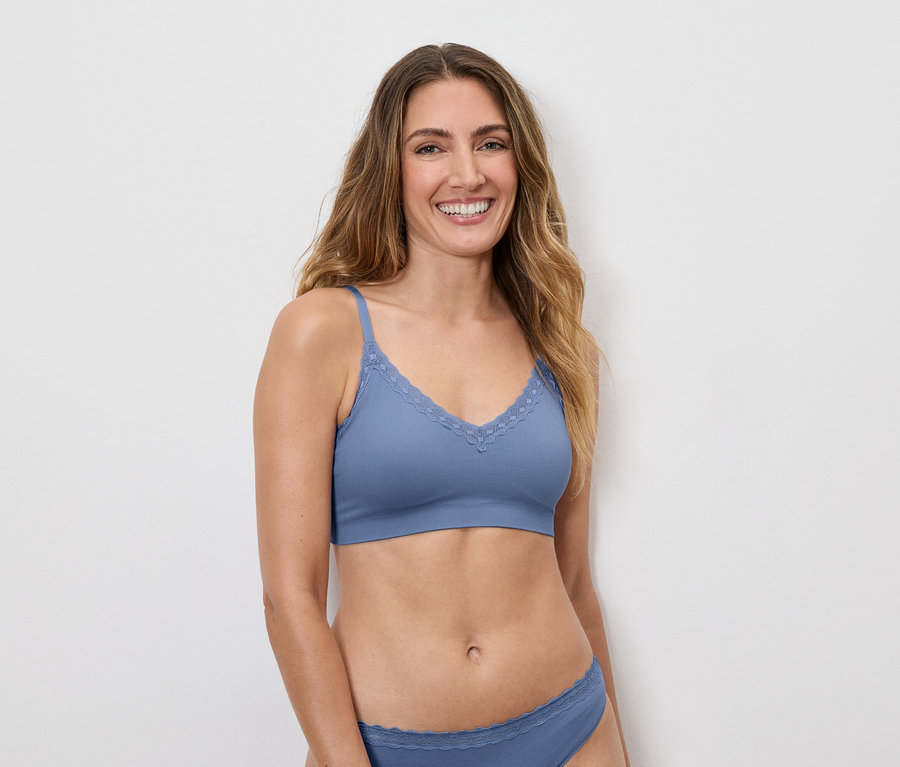 Une femme souriante aux longs cheveux bruns portant un soutien-gorge bleu avec des détails en dentelle et des sous-vêtements assortis sur un fond blanc.