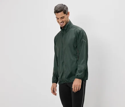 Un homme souriant portant une veste verte et des leggings noirs se tient devant un fond blanc.