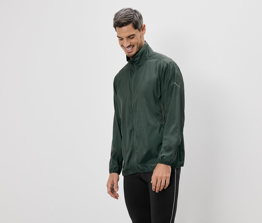 Un homme souriant portant une veste verte et des leggings noirs se tient devant un fond blanc.