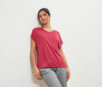 Jeune femme appuyée contre un mur, portant un t-shirt bordeaux et un pantalon gris.