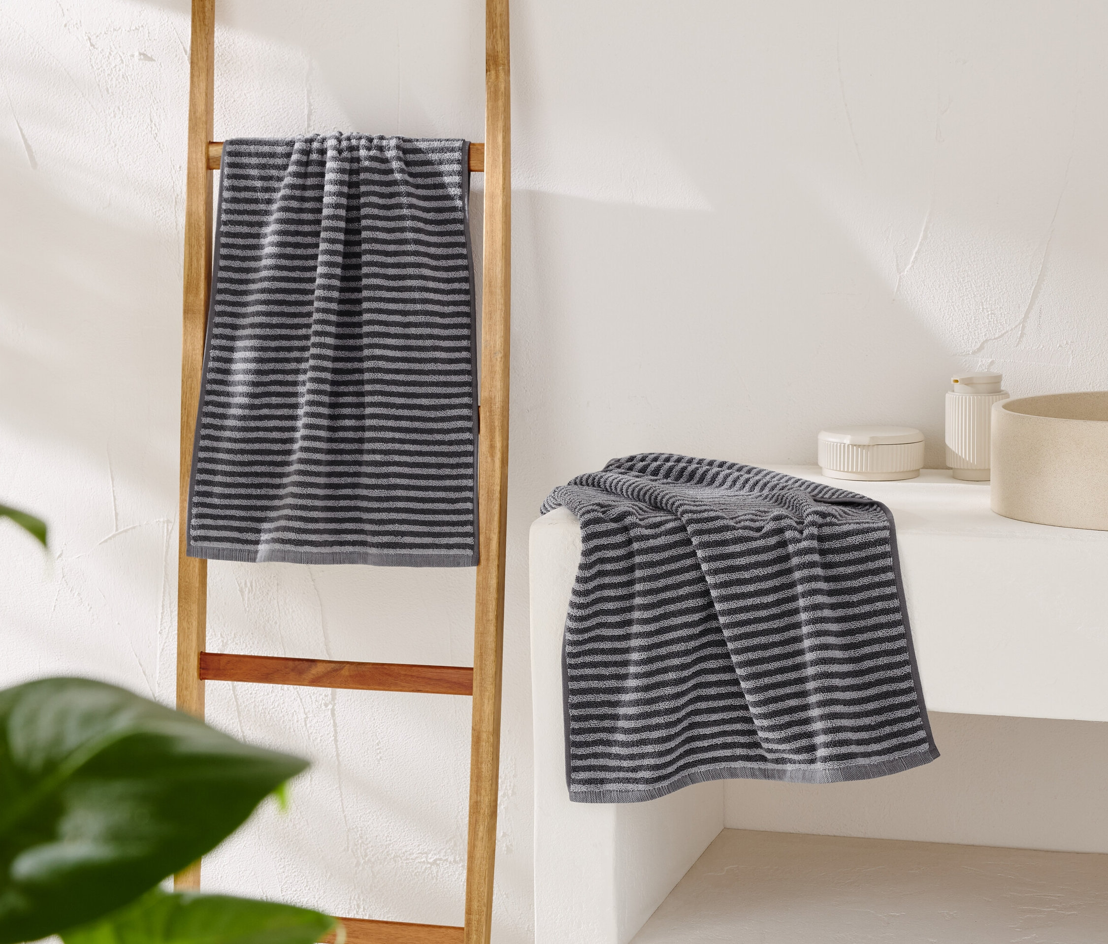 Une serviette grise rayée est accrochée à une échelle en bois, une autre est posée sur une coiffeuse blanche à côté d'un lavabo beige.