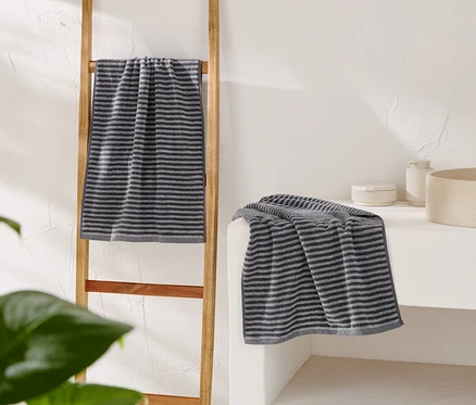 Une serviette grise rayée est accrochée à une échelle en bois, une autre est posée sur une coiffeuse blanche à côté d'un lavabo beige.