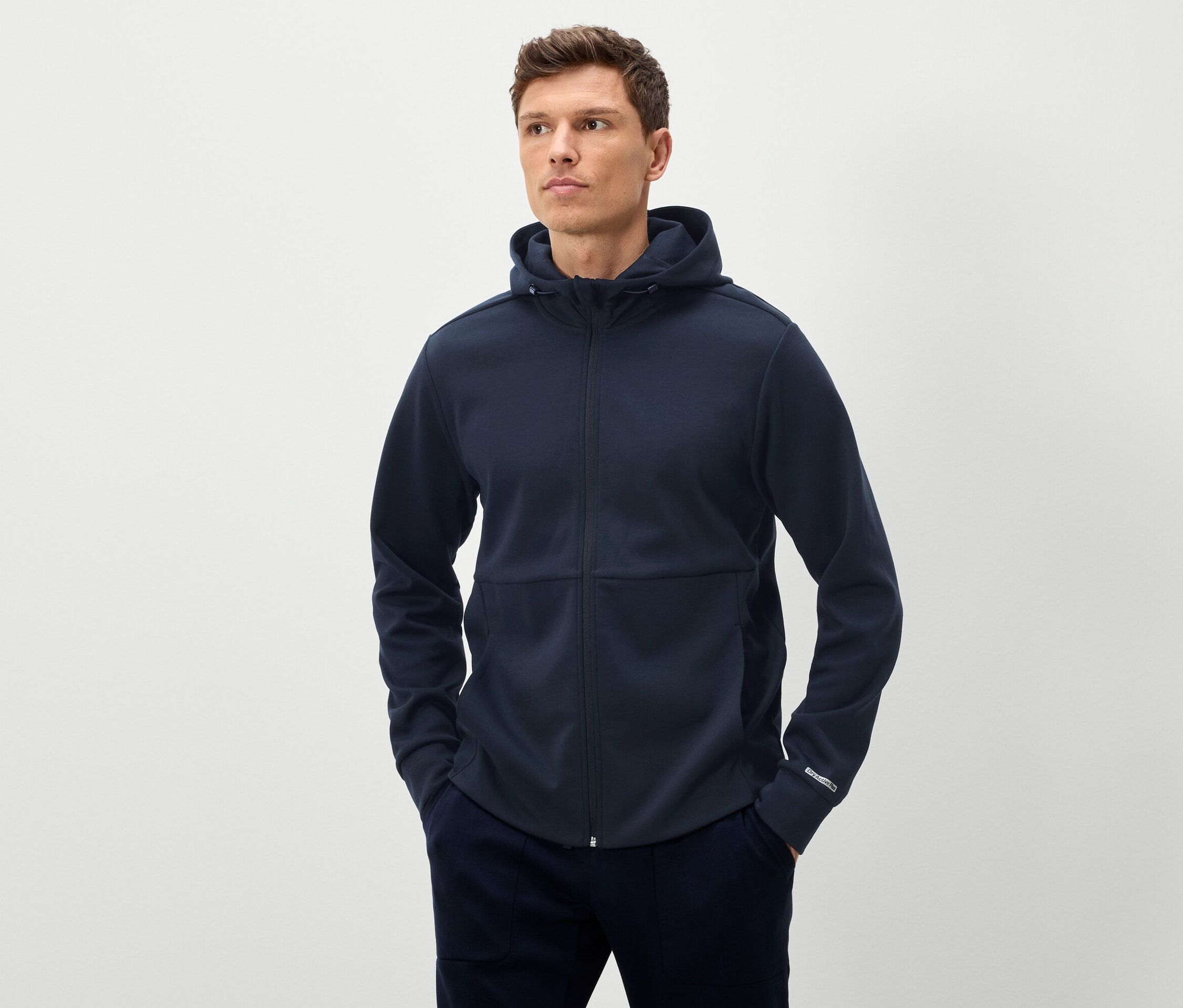 Homme posant en sweat-shirt à capuche bleu marine zippé sur toute la longueur et pantalon bleu marine, les deux mains dans les poches.