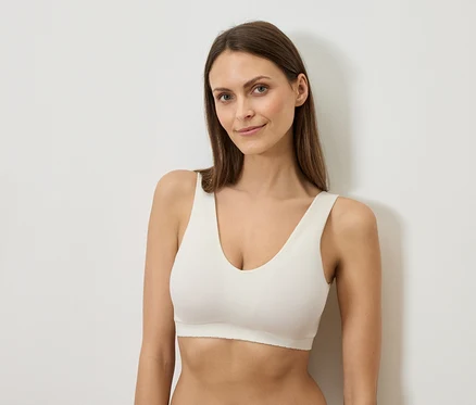 Femme aux longs cheveux bruns portant un soutien-gorge blanc appuyée contre un mur blanc.