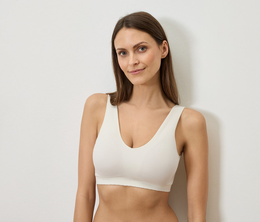 Femme aux longs cheveux bruns portant un soutien-gorge blanc appuyée contre un mur blanc.