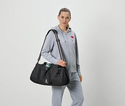 Femme portant un survêtement gris avec un cœur rouge et un pantalon de survêtement gris, tenant un sac de sport noir avec une bouteille d'eau.