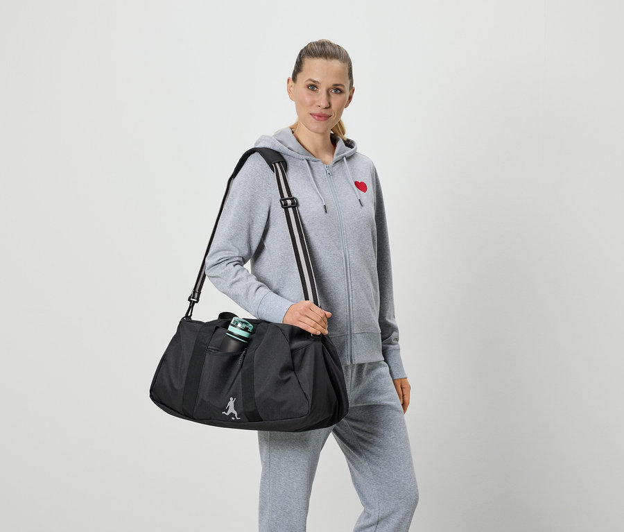Femme portant un survêtement gris avec un cœur rouge et un pantalon de survêtement gris, tenant un sac de sport noir avec une bouteille d'eau.