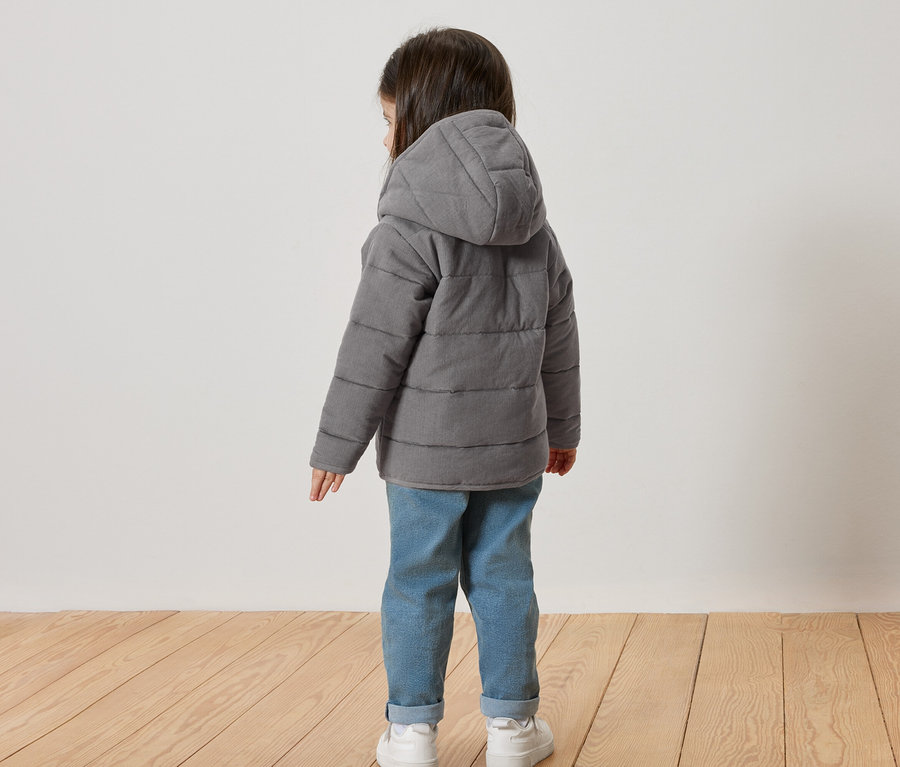 Enfant vu de dos, portant une veste matelassée grise à capuche, un jean et des baskets blanches.