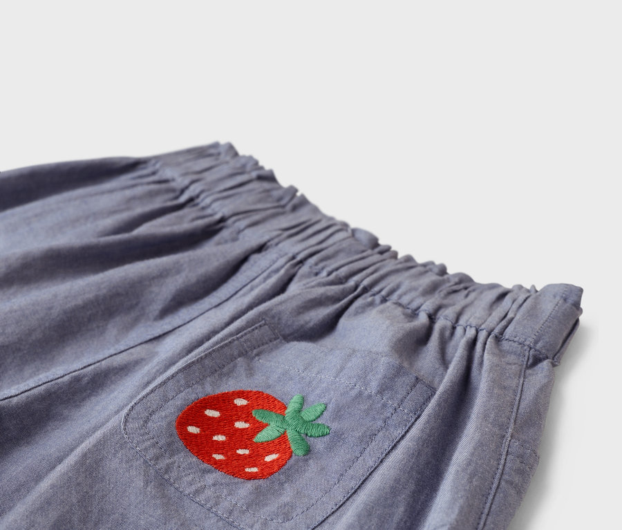 Gros plan d'un pantalon en tissu bleu avec une taille élastique et une poche plaquée avec une fraise rouge brodée.