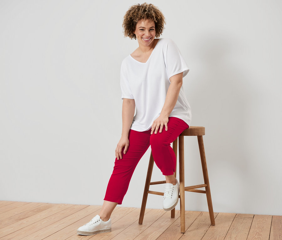 Femme aux cheveux bouclés assise sur un tabouret en bois, portant un t-shirt blanc et un pantalon rouge.
