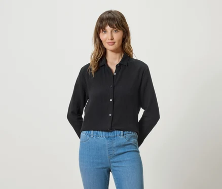 Une femme porte un chemisier noir à manches longues et un jean bleu sur un fond blanc.