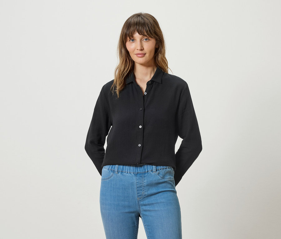 Une femme porte un chemisier noir à manches longues et un jean bleu sur un fond blanc.