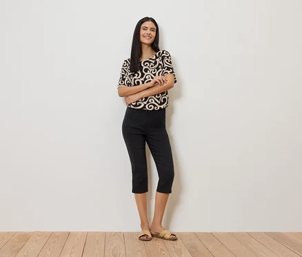 Femme portant un haut à motifs, un pantalon noir et des sandales devant un mur blanc.
