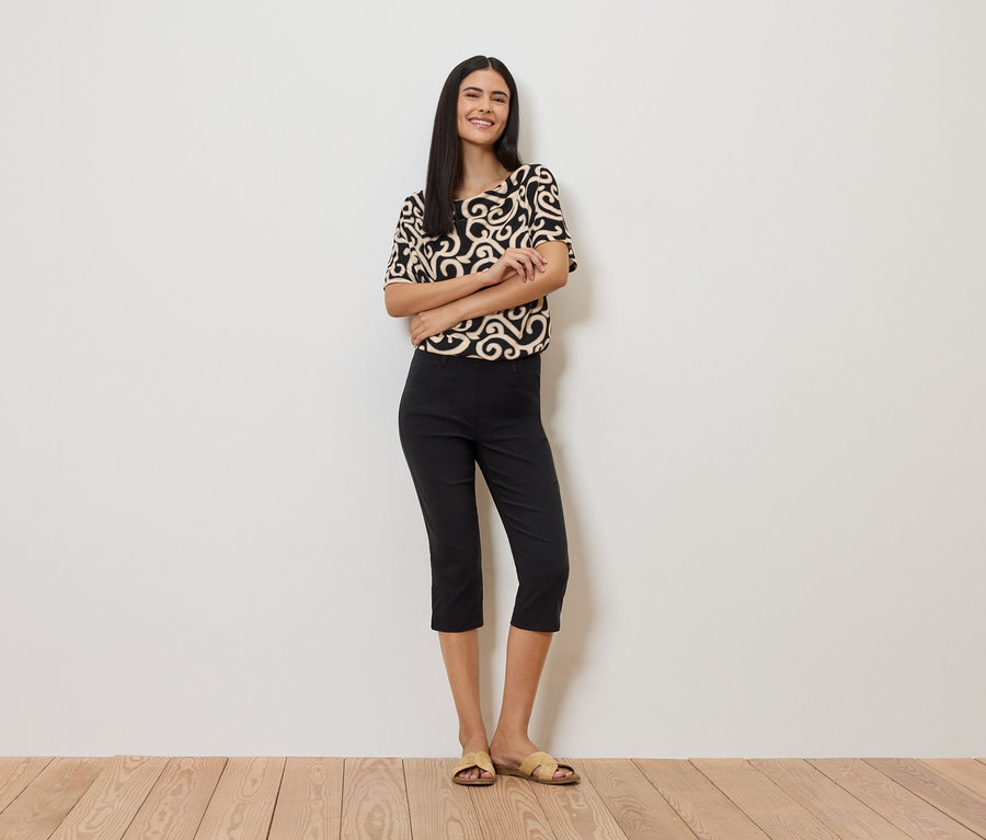 Femme portant un haut à motifs, un pantalon noir et des sandales devant un mur blanc.