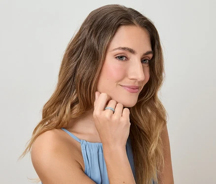 Une femme aux cheveux ondulés et au débardeur bleu touche son doigt orné d'une bague devant un fond clair.
