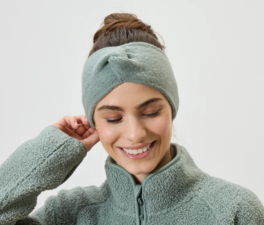 Une femme souriante, les cheveux relevés, portant un bandeau vert clair et une veste en polaire, touche son oreille.