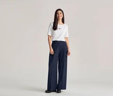 Une femme souriante aux longs cheveux foncés porte un t-shirt blanc avec trois cœurs rouges et un pantalon large bleu marine.