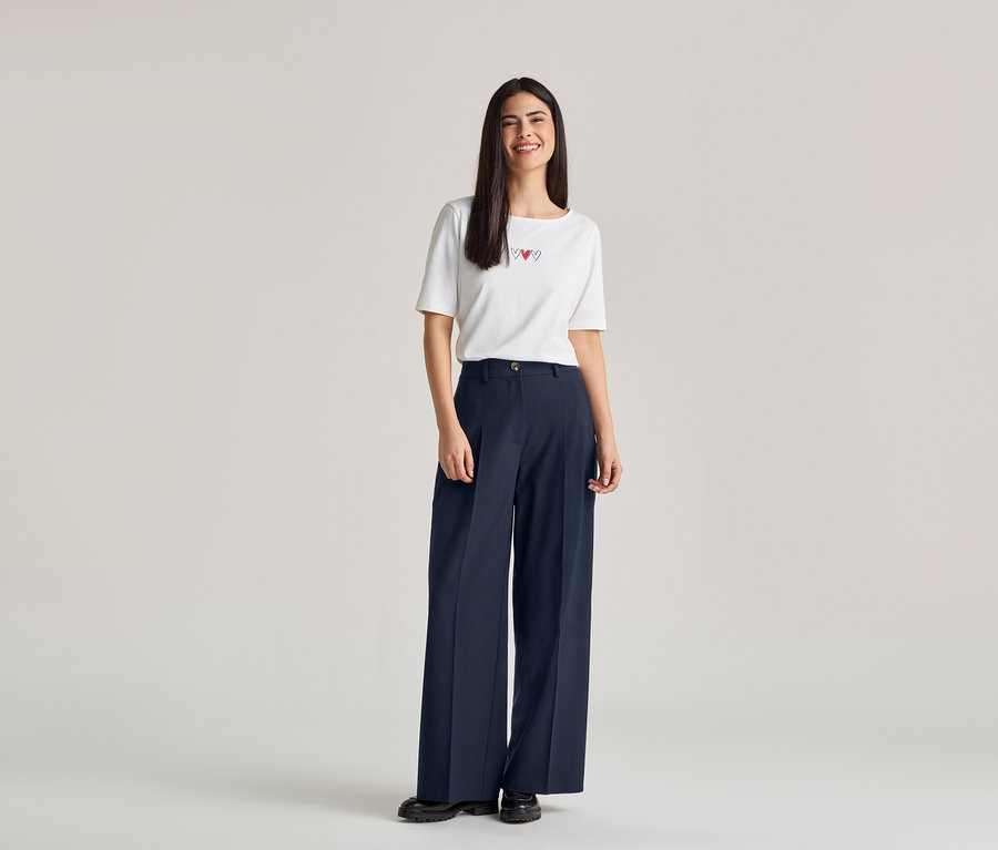 Une femme souriante aux longs cheveux foncés porte un t-shirt blanc avec trois cœurs rouges et un pantalon large bleu marine.