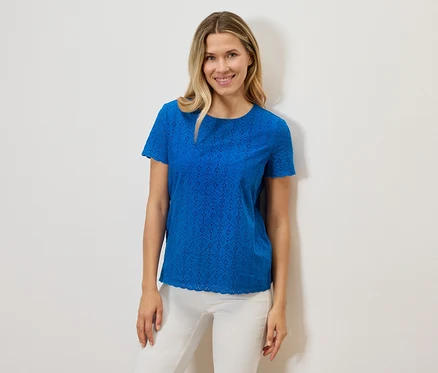 Une femme souriante porte un t-shirt bleu à manches courtes ajouré et un pantalon blanc sur un fond blanc.