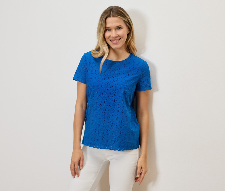 Une femme souriante porte un t-shirt bleu à manches courtes ajouré et un pantalon blanc sur un fond blanc.