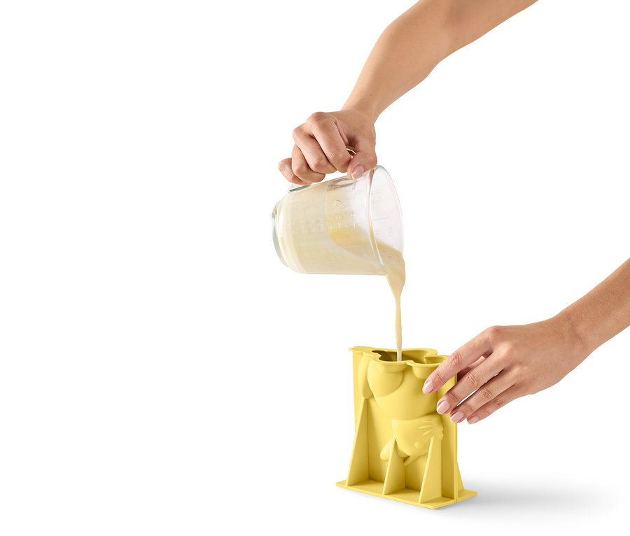 Une personne verse de la pâte d'un verre doseur dans un moule en silicone jaune en forme de chat.