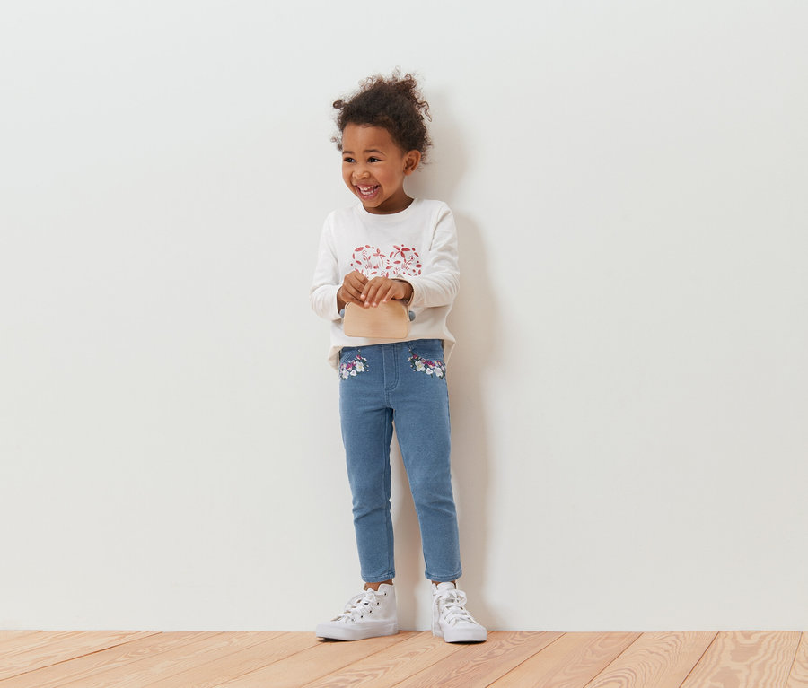 Fillette souriante aux cheveux bouclés tenant un petit sac beige, portant un haut à manches longues et un jean bleu avec des broderies florales, debout devant un mur blanc.