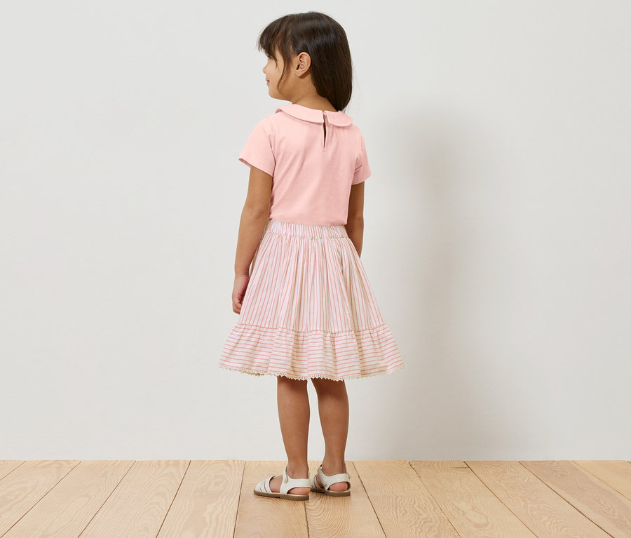 Une fille se tient dos à la caméra, portant un haut rose, une jupe rayée rose et blanche et des sandales blanches.