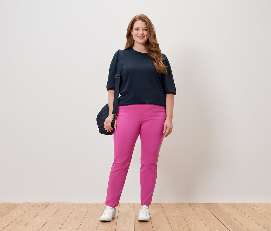 Femme debout en haut bleu marine et pantalon rose avec un sac à bandoulière devant un mur blanc.