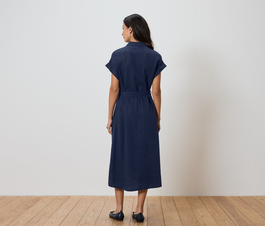 Une femme vue de dos portant une robe-chemise bleu marine avec une ceinture et des ballerines sur un fond blanc.