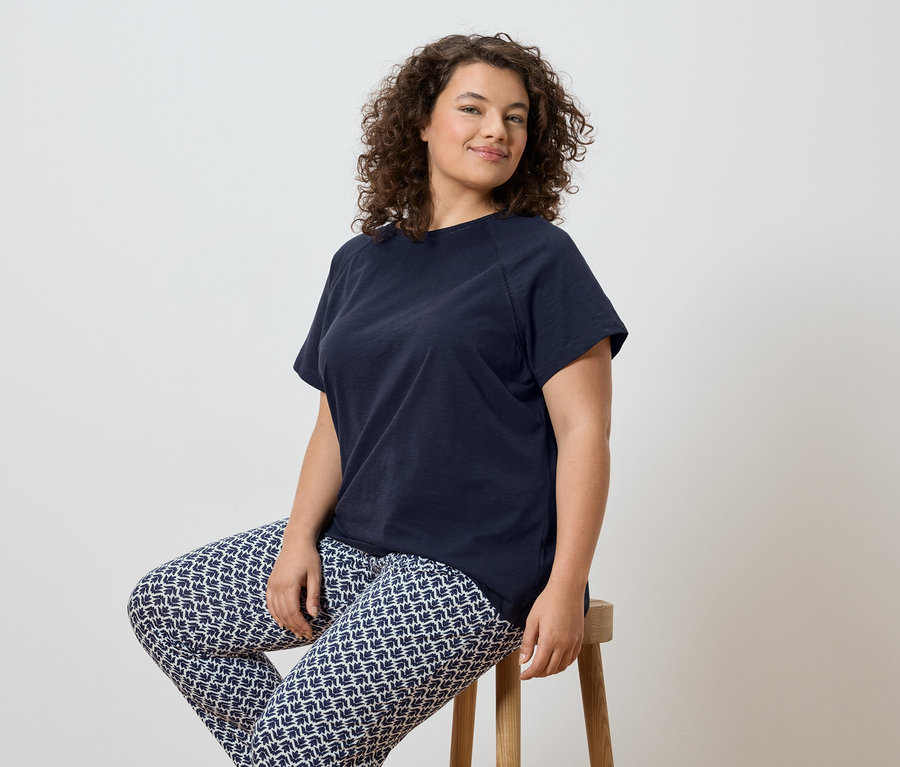 Une femme aux cheveux bouclés est assise sur un tabouret, portant un t-shirt bleu marine et un legging à motifs.